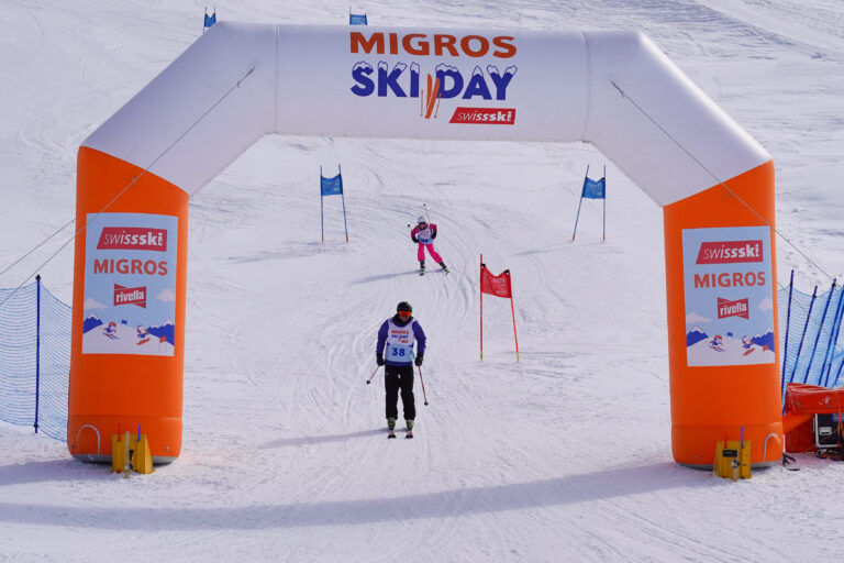 Skifahrer unter Migros Skiday Torbogen mit Rivella Werbung.