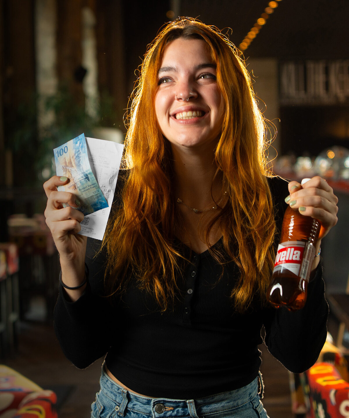 Junge Frau hält Rivella Rot Flasche und Geldschein, lächelnd in Café.