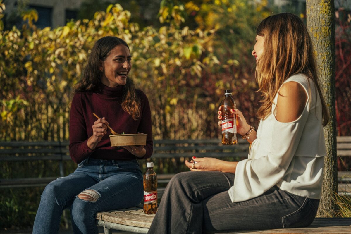 Zwei Frauen geniessen Rivella Rot in herbstlicher Parklandschaft.