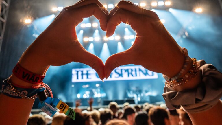 Zwei Hände formen Herz vor bunter Konzertbühne mit Scheinwerfern.