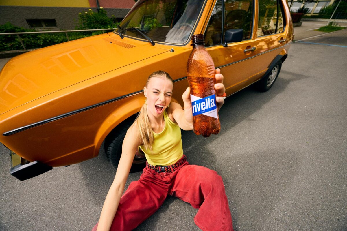 Frau hält Rivella Zero Flasche vor orangefarbenem Auto.