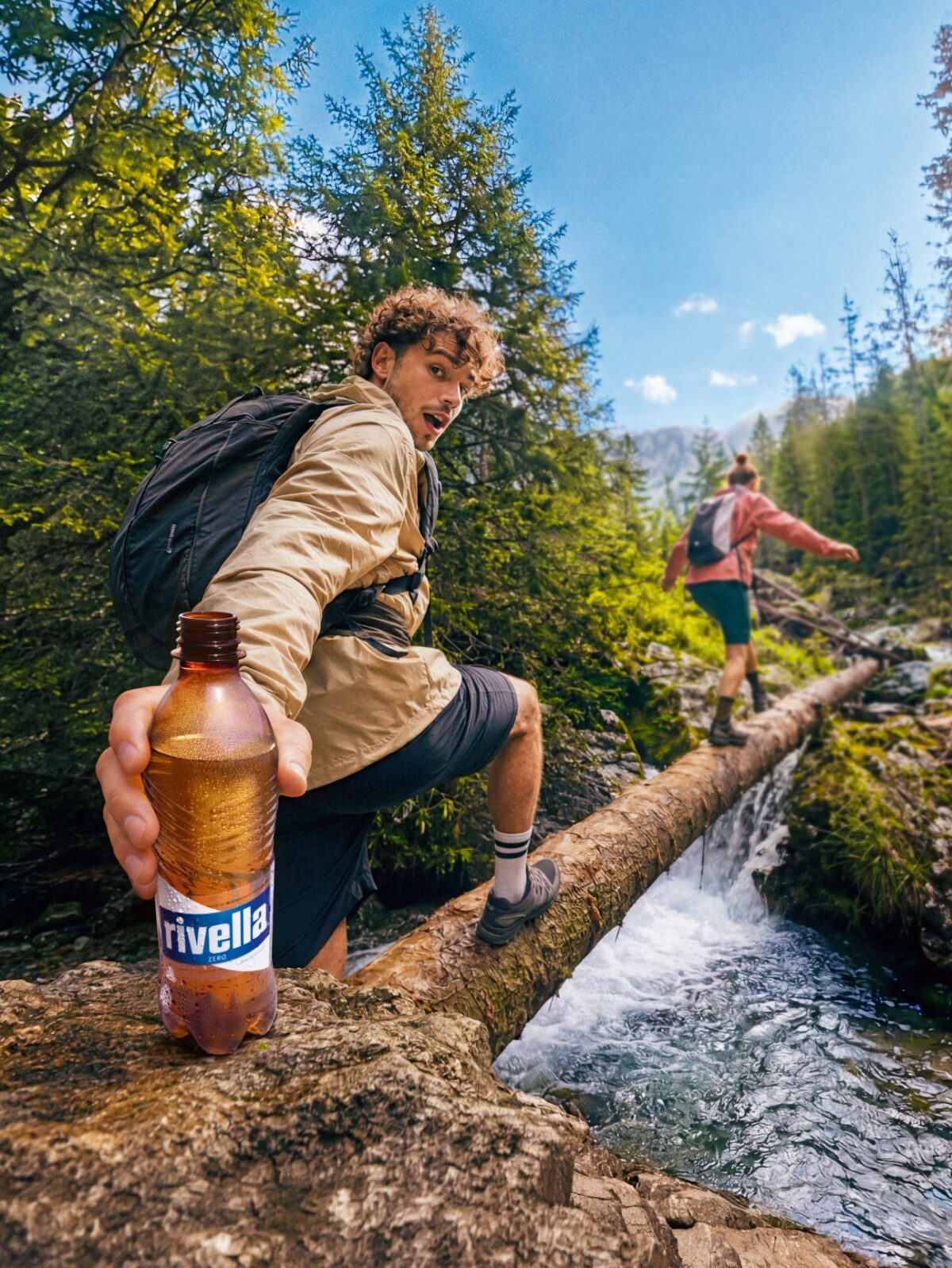 Wanderer zeigt Rivella Zero Flasche beim Balancieren über Bach im Wald.
